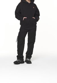 Persona con sudadera negra con un pequeño logo en el pecho, pantalones cargo negros con bolsillos laterales y zapatillas negras con cordones blancos, de pie con las manos en los bolsillos.