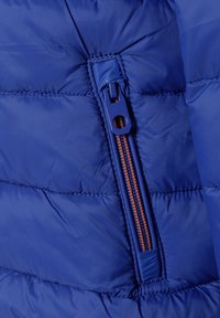 Podrobnost modre napihnjene jakne, ki vključuje zadrgo z oranžnimi poudarki, kviltano teksturo in gladko površino.