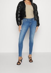 Chaqueta puffer negra corta, top beige, vaqueros pitillo azules y zapatos de tacón negros con tiras. Texturas suaves y ajuste ceñido.