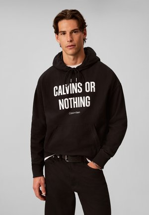 Jeune homme portant un sweat à capuche noir avec le texte "CALVINS OR NOTHING" et un pantalon noir sur un fond gris uni.