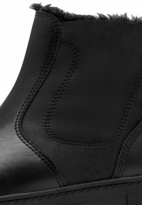 Schwarze Lederstiefelette mit einem kuscheligen, pelzigen oberen Rand, glatter Oberfläche und kontrastierenden, genähten Details entlang der Seiten und Sohle.