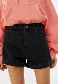 Shorts de mezclilla negros de cintura alta, con dobladillo enrollado y bolsillos frontales, combinados con una sudadera con capucha rosa.
