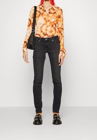 En långärmad skjorta i orange och brunt mönster, svarta tajta jeans och glänsande svarta skor. Bär en liten svart väska, stående mot en neutral bakgrund.