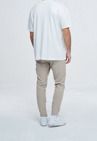 Muž v bílé košili s krátkým rukávem, béžových kalhotách a bílých teniskách, stojící proti jednobarevnému světlému šedému pozadí, viděný zezadu.