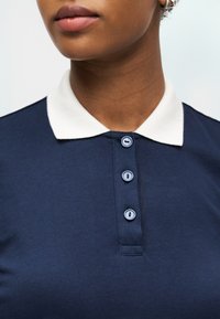 Femme portant un polo bleu marine avec un col blanc et trois boutons bleu clair, gros plan montrant les lèvres, le cou et l'oreille avec une boucle d'oreille.