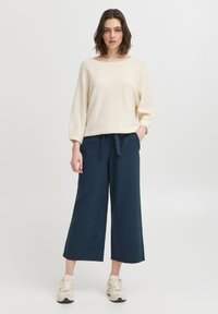 Un pull en tricot crémeux avec une texture, à manches larges, associé à un pantalon large bleu marine avec cordon de serrage. Les chaussures sont des baskets de couleur claire.