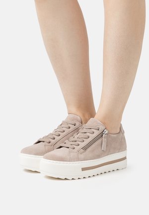 Pieds portant des baskets compensées en daim beige avec des fermetures éclair latérales et des semelles blanches rainurées sur un fond uni.