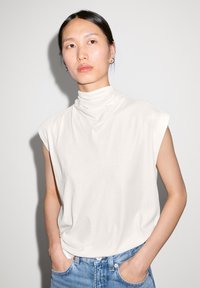 White, sleeveless turtleneck top with draped neck, made of soft fabric, paired with light blue denim jeans, hands in pockets.