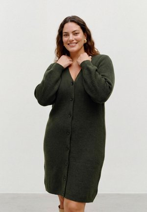 Une femme souriante porte une robe en maille vert foncé, à manches longues et boutonnée sur le devant, sur un fond uni et clair.