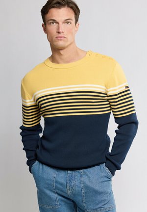Homme portant un pull rayé jaune et marine avec des boutons sur les épaules et un jean bleu, debout les mains dans les poches devant un fond gris.