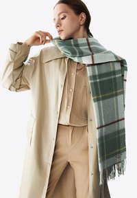 Sciarpa a quadri verde e grigia realizzata in tessuto morbido, con bordi frangiati. Indossata con una camicia beige con bottoni e un cappotto beige chiaro.