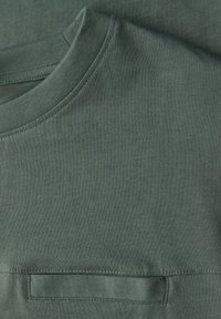 Zelené bavlněné tričko s kulatým límečkem, hladkou texturou a malou obdélníkovou nášivkou. Jednoduchý design bez viditelných vzorů nebo zdobení.