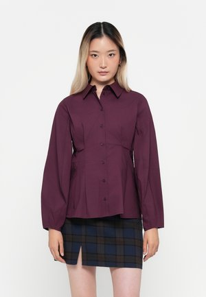Chemise boutonnée maroon à manches longues, design devant plissé, associée à une mini jupe à carreaux sombre avec une fente sur le côté.