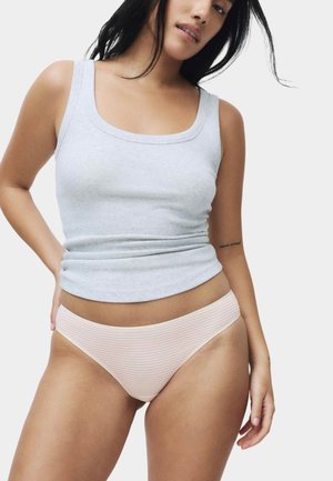 Femme portant un débardeur gris clair et un sous-vêtement style bikini à rayures rose clair, debout avec une main sur la hanche.