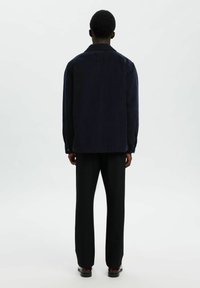 Persona de pie con la espalda hacia el espectador, vistiendo una chaqueta de pana color azul marino, pantalones negros y zapatos oscuros pulidos, contra un fondo liso.