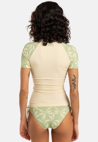 Conjunto de camiseta de protección y bikini en color beige claro y verde. La camiseta de protección tiene mangas cortas y un patrón de hojas de palma; el bikini tiene lazos ajustables.