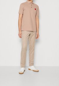 Beżowa koszulka polo z krótkim rękawem, zapinana na trzy guziki, z małym czerwonym logo, połączona z jasnymi, w kratkę beżowymi spodniami oraz białymi trampkami na gumowej podeszwie.