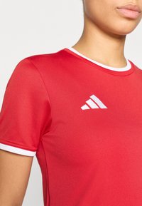 Κόκκινο αθλητικό μπλουζάκι  με κοντά μανίκια, στρογγυλή λαιμόκοψη, λευκή τρέσα στον γιακά και κεντητό λευκό λογότυπο στο στήθος. Ομαλή υφή υφάσματος.