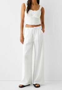 Biały, krótki top na ramionkach z marszczeniami i falbaniastymi brzegami, zestawiony z szerokimi, wysoko taliowanymi spodniami z białego lnu i płaskimi czarnymi sandałami.