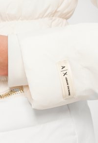 Manica di giacca a vento bianca con polsino a costine. Presenta un'etichetta in tessuto con "A|X ARMANI EXCHANGE" in beige, cucita in colori a contrasto.