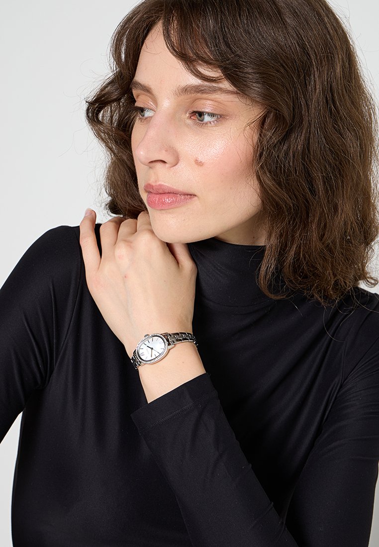 Mujer con cabello castaño ondulado vistiendo un suéter negro de cuello alto y un reloj de plata en la muñeca, mirando hacia la izquierda con la mano cerca del cuello.