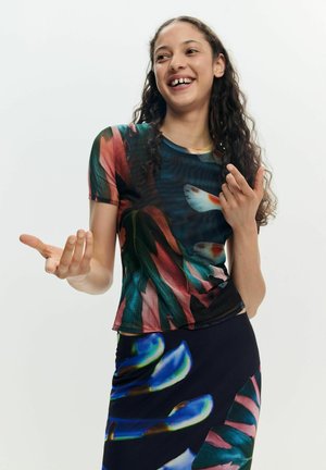 Tânără cu păr lung și creț, purtând un top și o fustă colorate, cu model, zâmbind și gesticulând cu mâinile pe un fundal simplu.