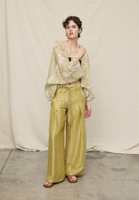 Jeune personne debout portant un pantalon ample vert olive, un chemisier beige à motifs, et des sandales marron devant un fond en tissu beige uni.