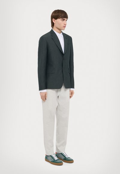 Blazer gris foncé à deux boutons, associé à une chemise blanche et un pantalon gris clair. Les chaussures sont en daim vert avec des lacets blancs.
