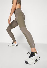 Leggings verde oliva con pannelli testurizzati, vita alta e dettaglio con logo viola. Abbinati a scarpe da ginnastica nere e bianche.