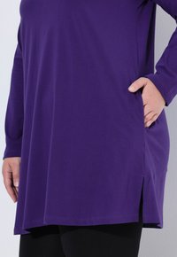 Fioletowa tunika z długim rękawem, charakteryzująca się luźnym krojem, bocznymi rozcięciami oraz miękkim bawełnianym materiałem. Zestawiona z czarnymi legginsami dla kontrastu.