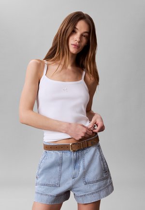 Młoda kobieta poprawiająca biały top na ramiączkach, ubrana w jasne szorty jeansowe z brązowym paskiem, stojąca na jednolitym szarym tle.