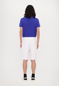 Blaues Poloshirt mit kurzen Ärmeln und schlichtem Design, kombiniert mit weißen knielangen Shorts. Die Person trägt schwarze Socken und weiße Sneakers.