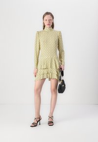 Rochie galbenă cu un model, cu guler înalt, mâneci lungi și hem în straturi. Asortată cu o geantă neagră cu umplutură și sandale negre cu curele.