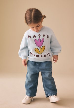 Peuter met bruin haar in vlechtjes, draagend een lichtblauwe trui met "Happy Moments" en een bloemenprint, blauwe spijkerbroek en witte sneakers.