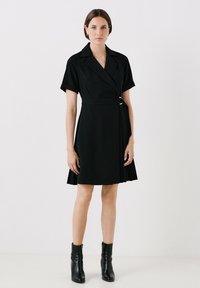 Robe noire à manches courtes, avec une taille cintrée mettant en avant une boucle en argent, et une jupe plissée. Portée avec des bottines noires.