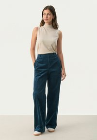 Beige mouwloos topje gecombineerd met high-waisted, teal corduroy broek. De broek heeft een wijdrecht ontwerp. Model draagt lichtgekleurde schoenen.