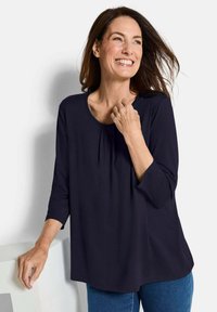 Marineblaues Blusenshirt mit Dreiviertelärmel, rundem Ausschnitt und Faltendetail. Weicher Stoff mit lockerem Sitz und abgerundetem Saum.