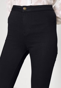 Pantalones de mezclilla negros de talle alto con un botón dorado, que presentan una textura suave y bolsillos laterales, diseñados para una silueta ajustada.