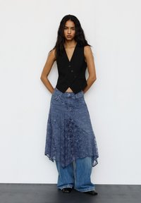 Egy nő fekete ujjtalan mellényben, átlátszó kék csipke aszimmetrikus szoknyában, széles lábú farmert viselve, fekete hegyes cipőben áll egy fehér falnak támaszkodva.