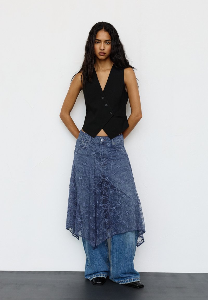 Egy nő fekete ujjtalan mellényben, átlátszó kék csipke aszimmetrikus szoknyában, széles lábú farmert viselve, fekete hegyes cipőben áll egy fehér falnak támaszkodva.