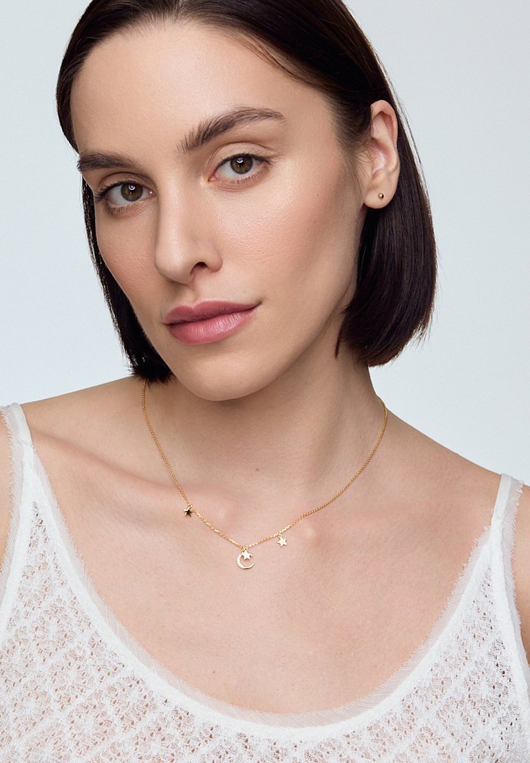 Złoty naszyjnik z zawieszkami w kształcie półksiężyca i gwiazdy na delikatnym łańcuszku, noszony na białej, teksturowanej bluzce z cienkimi ramiączkami.