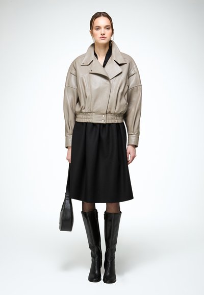 Femme portant une veste en cuir beige, une robe midi noire, des bottes noires montantes, tenant un sac à main noir, debout sur un fond blanc.