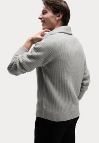 Suéter de canalé gris claro con cuello de chal y mangas largas, que presenta un diseño de punto texturizado. El modelo está sonriendo y ajustando el cuello.