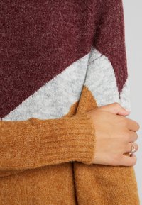 Färgblockerad tröja med vinröda, grå och bärnstensfärgade nyanser. Mjuk, strukturerad stickning med ribbade ärmslut och ett geometriskt mönster.