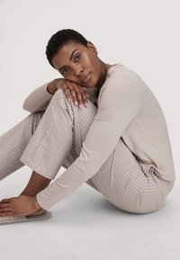 Eine Frau mit kurzen Haaren in einem beigen Langarmshirt und gestreiften Hosen sitzt zusammengekauert auf dem Boden und ruht ihren Kopf auf den Knien.