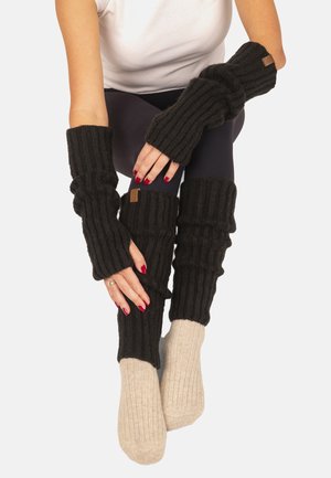 Person iført sorte, ribstrikkede fingerløse handsker og matchende leggvarmere over beige ribstrikkede sokker, siddende med hænderne hvilende på benene.