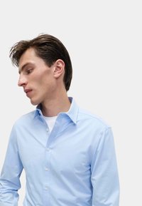 Svetle modrá košile s dlouhým rukávem, s límcem na knoflíky, slim fit, hladká textura a bílé knoflíky, nošena přes bílý tričko.
