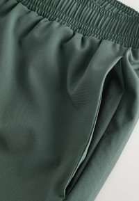 Pantalon à taille élastique vert foncé avec poche latérale, montrant la texture du tissu et les détails des coutures.