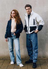 To personer poserer foran en ensfarvet væg. Kvinden har en marineblå jakke, en beige hættetrøje og lyse jeans på, mens manden har en hvid og sort fleecejakke samt mørke jeans.