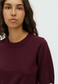 Pull en maille bordeaux avec un col rond, des manches courtes et une texture lisse. Présente un design ajusté et des détails de couture subtils.